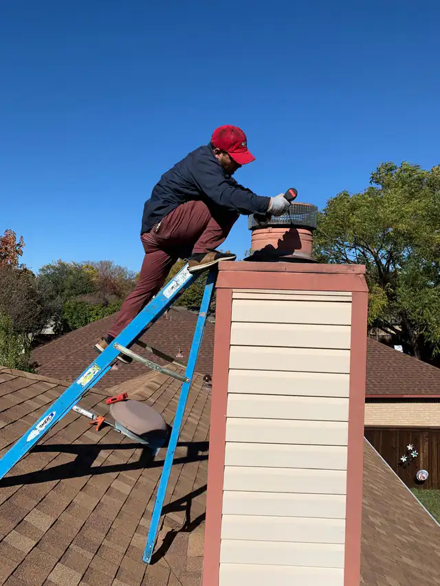 A technician fastening a stainless mesh rain cap onto a clay flue at the top of a sided chimney chase