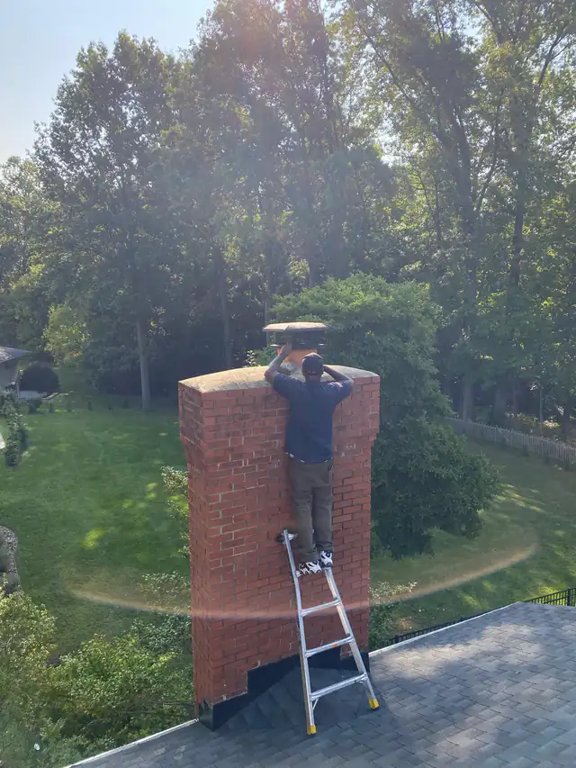 A technician on a ladder set against a tall brick chimney, fitting a rain cap at the top