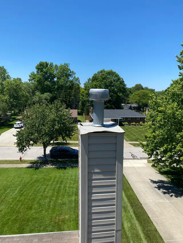The top of a sided chimney chase before replacement, with a distorted pale metal cover and a round vent above it