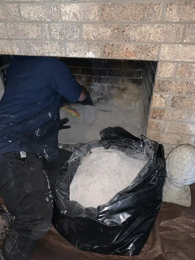 A technician working inside an open firebox with a bag of removed material beside the hearth on protective sheeting