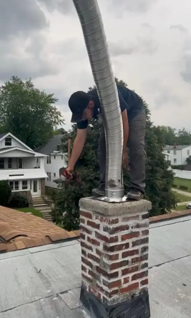 A technician on a flat roof feeding a flexible stainless steel chimney liner down into a brick chimney