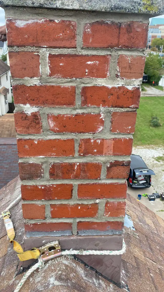 A freshly repointed brick chimney above a shingle roof, with a rope-and-harness roof anchor and sealed flashing at its base