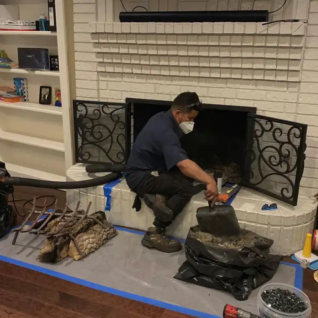 A technician in a respirator sweeping a firebox, with drop cloths taped to the floor, tools laid out on a mat and a vacuum hose run to the hearth