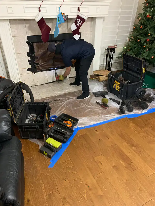 A technician working at a taped-off fireplace opening, with floor protection and chimney brushes beside the hearth