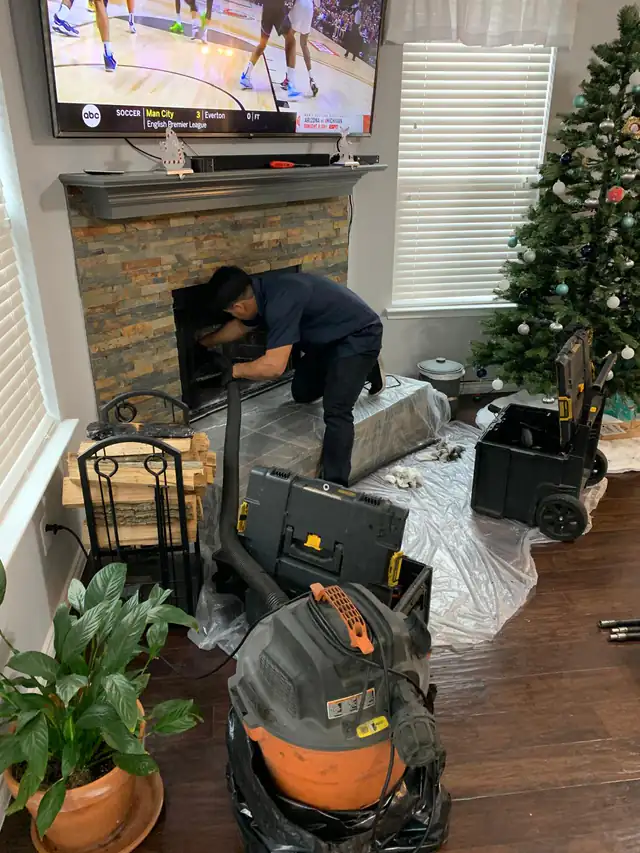 A technician reaching into a stone-front fireplace, with protective floor covering and a vacuum beside the hearth